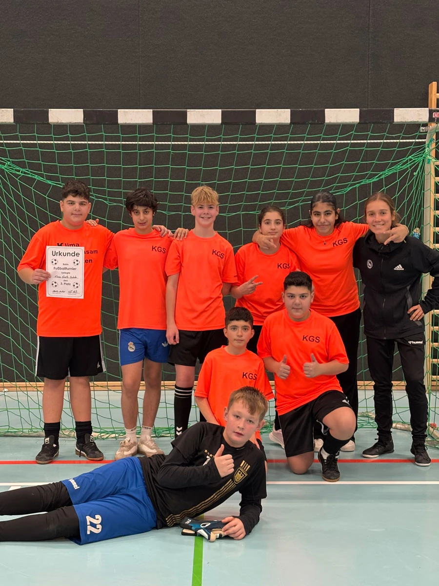 5 Kinder plus Trainerin stehen im Hintergrund, zwei knien davor, ein Kind liegt vorne davor